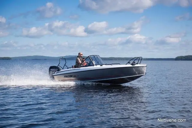 A man drives a Falcon boat across a lake, creating splashes in the water under a clear blue sky and distant greenery.